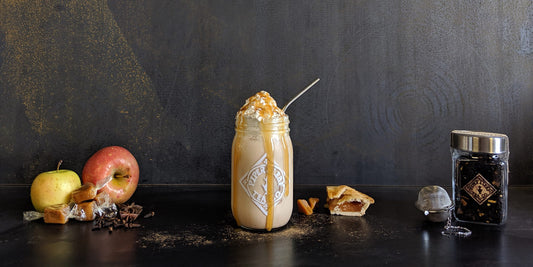 A pint jar filled with tea and topped with whipped cream and a caramel drizzle. Around it are apples, caramel candies, a slice of pie, and a jar of loose leaf