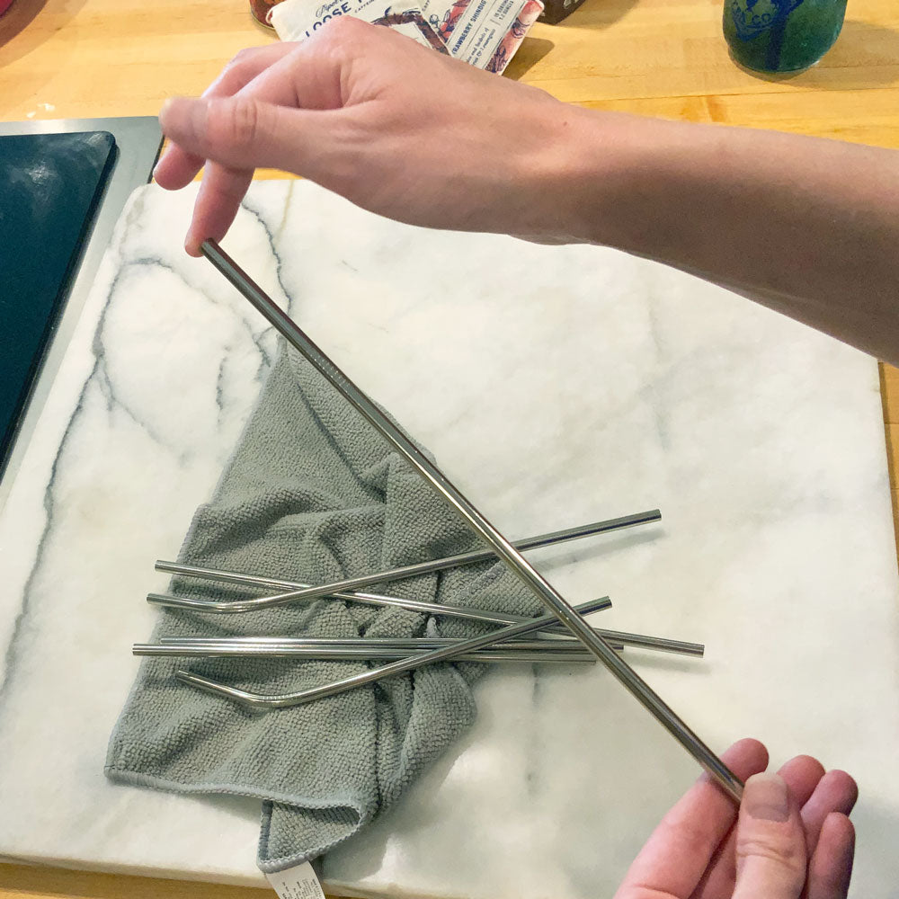 Hands holding a shiny metal straw up to the camera. Behind it on the counter are more straws laid on top of a grey towel.
