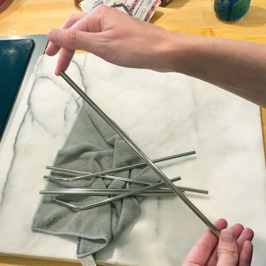 Hands holding a shiny metal straw up to the camera. Behind it on the counter are more straws laid on top of a grey towel.