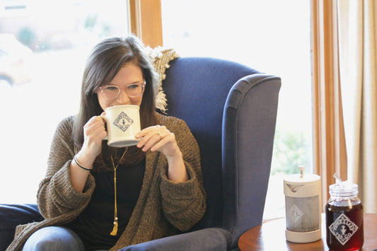 A woman in glasses sips hot tea from her Piper & Leaf mug beside her freshly brewed concentrated tea and her Piper Press