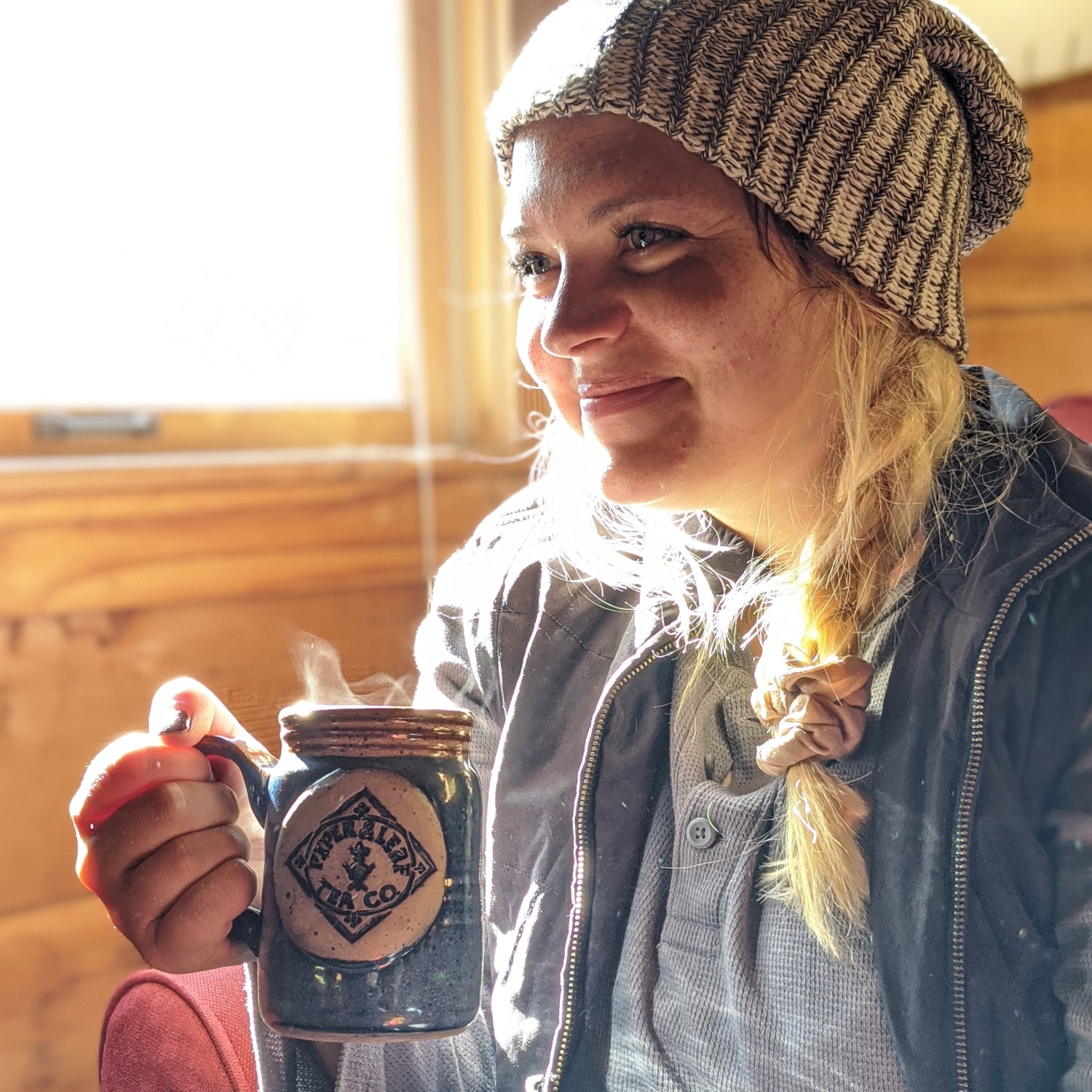 Person wearing a knit hat holds a steaming mug inside a wooden room, smiling softly while looking to the side.
