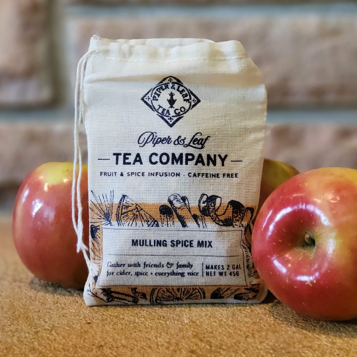 A cloth bag labeled "Piper & Leaf Tea Company Mulling Spice Mix" sits between two red apples on a brown surface.