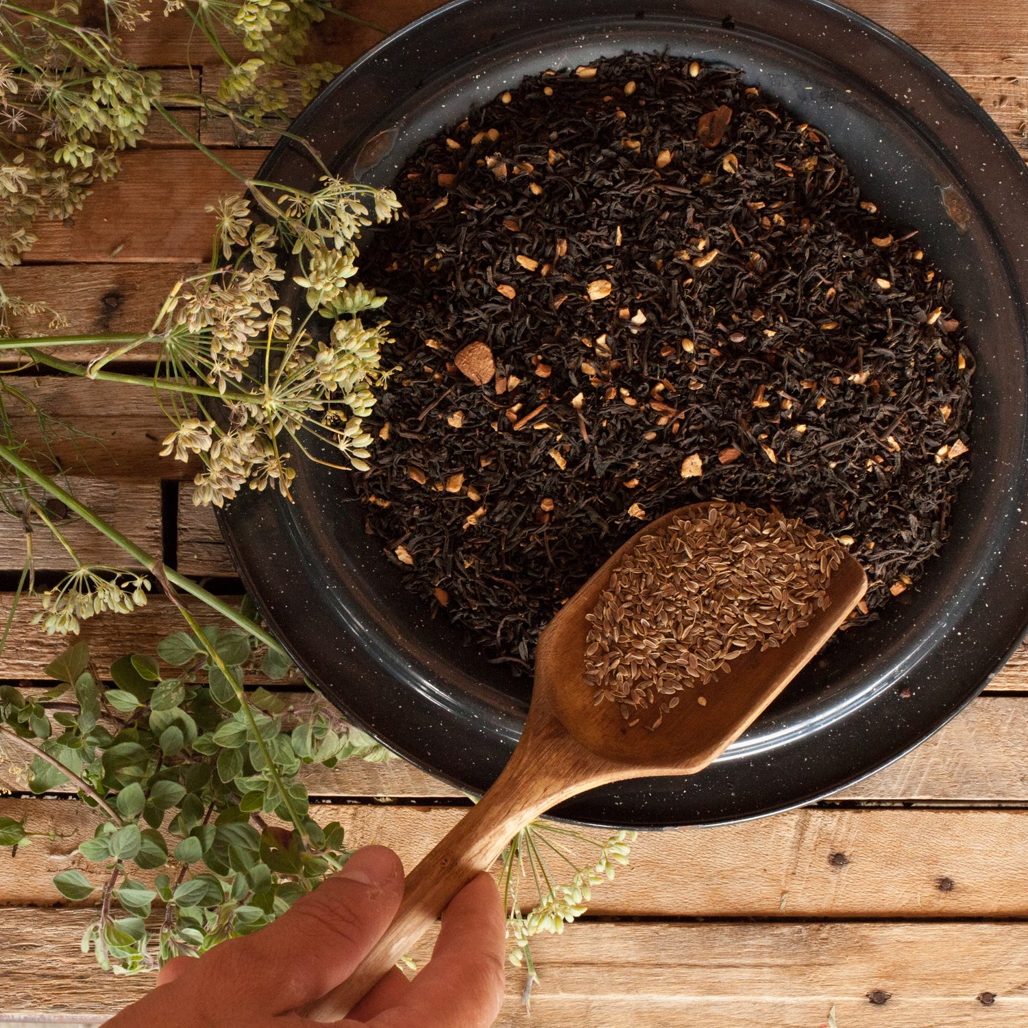 A hand holding a wooden spoon with cumin seeds over a bowl of black tea leaves on a wooden surface.
