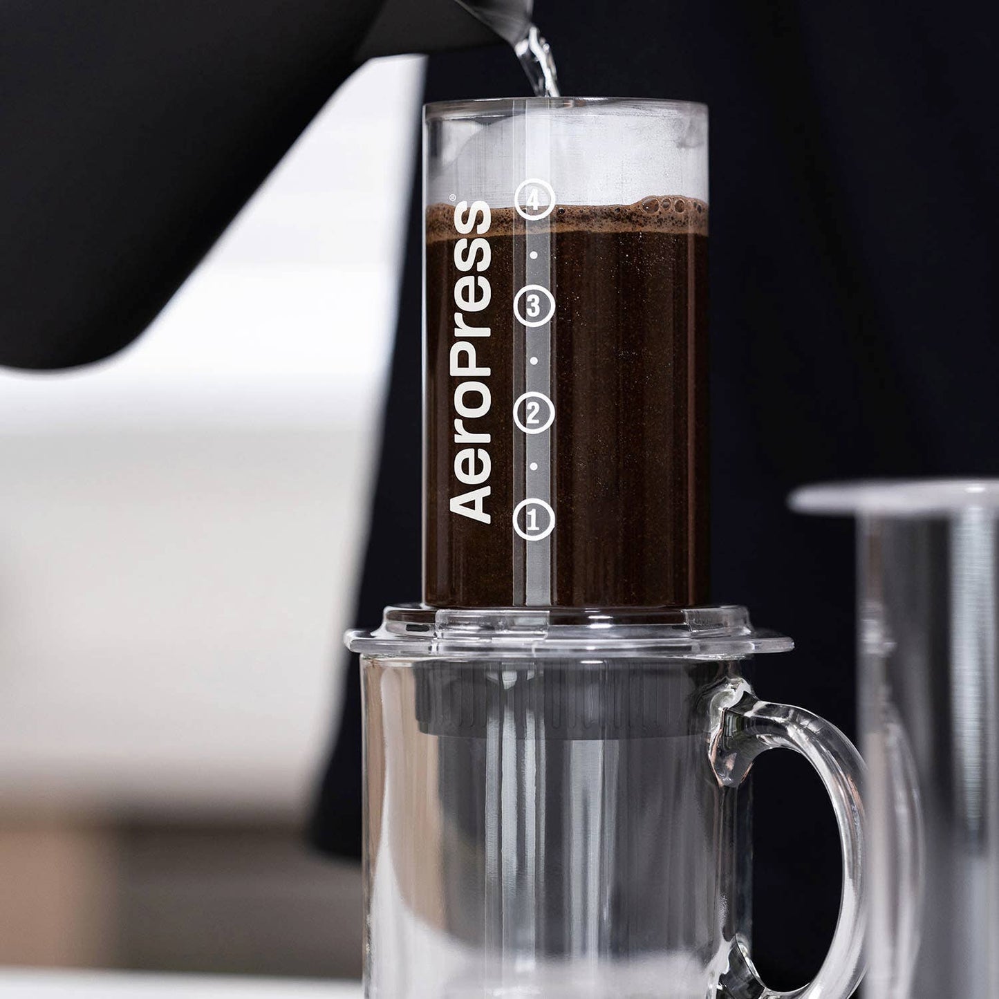 Hot water is poured into an AeroPress coffee maker filled with ground coffee and fitted with AeroPress Paper Micro-Filters by AeroPress, positioned above a clear glass mug.
