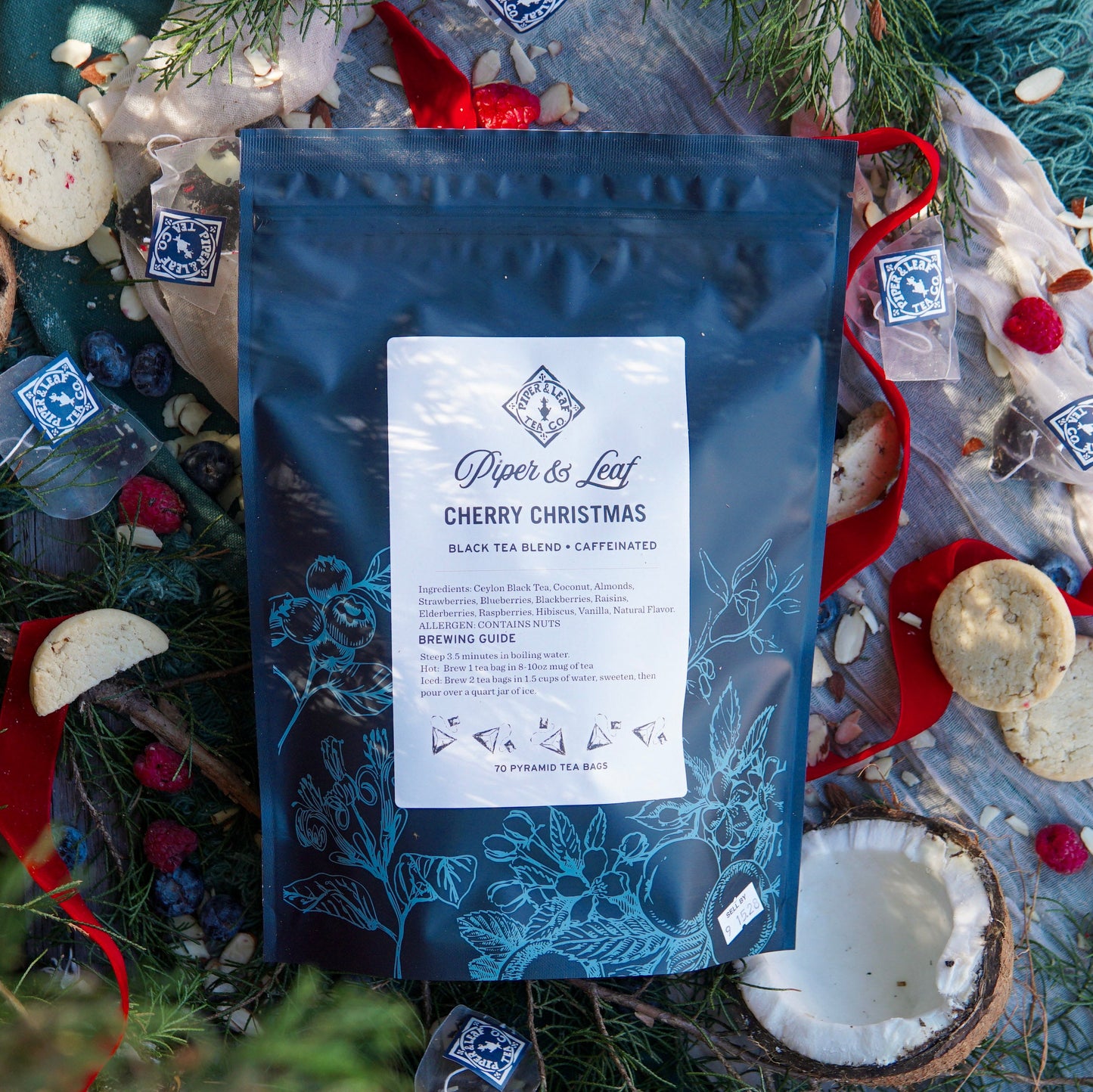 A bag of Piper & Leaf Tea Co. Cherry Christmas Bulk Sachets (70ct tea bags) rests on a festive surface surrounded by cookies, ribbons, greenery, and loose tea pyramids.