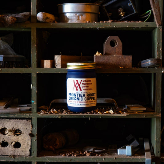 A 5oz jar of Walling Mtn. Coffee’s Frontier Roast sits on a shelf in a cluttered, dimly lit storage cabinet filled with tools and hardware, its rich aroma hinting at a dark roast crafted by Walling Mtn. Coffee.
