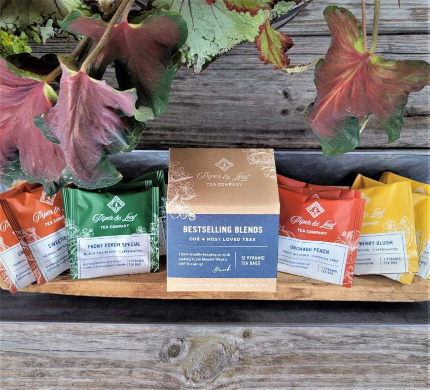 A box labeled "Bestselling Blends- Box of 12 Tea Bags" by Piper & Leaf Tea Co. is surrounded by colorful tea packets, featuring a captivating black tea blend, on a wooden surface with leafy decor.
