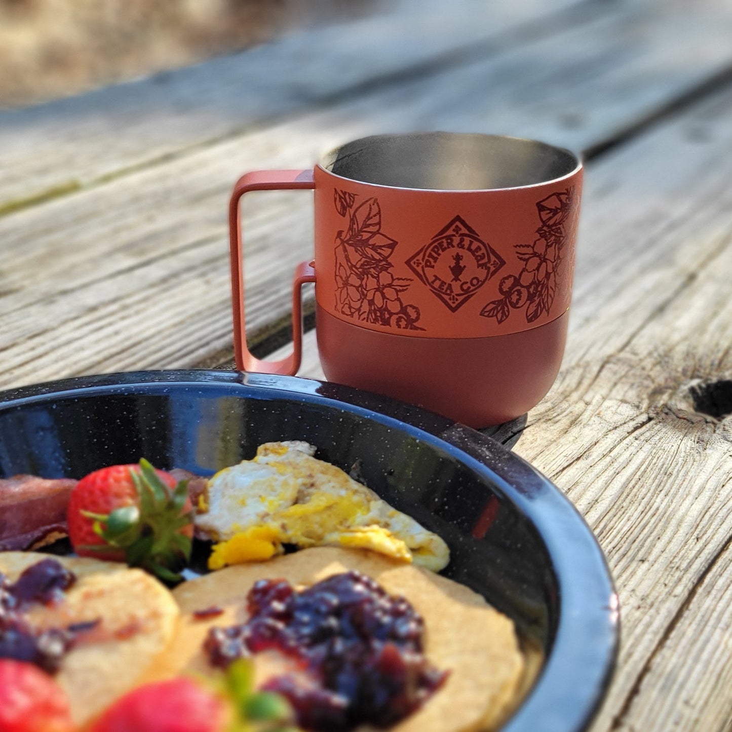 A plate of pancakes with jam, eggs, bacon, and strawberries sits on a wooden table beside the 14oz P&L Rise Mug by Klean Kanteen from Piper & Leaf Tea Co.—the ideal companion for tea lovers enjoying breakfast outdoors.