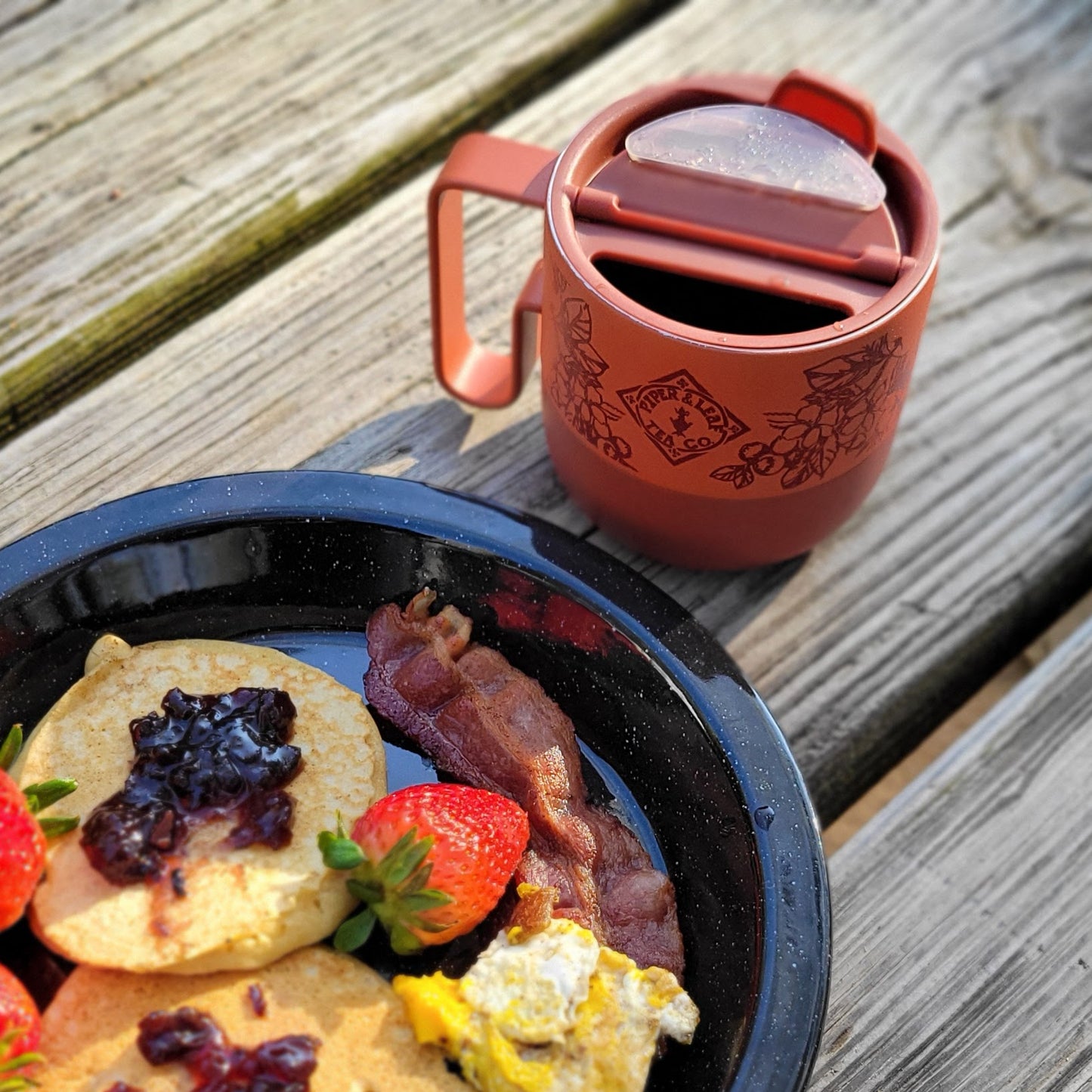 A plate with pancakes, jam, strawberries, bacon, and eggs sits on a wooden table beside a Piper & Leaf Tea Co. P&L 14oz Rise Mug from Klean Kanteen—a red recycled stainless steel mug with lid—ideal for tea lovers.