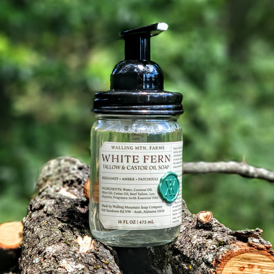 A 12oz glass pump bottle of Walling Mountain Farms White Fern Tallow & Castor Oil Foaming Hand Soap rests on a tree branch, with a blurred green forest in the background.