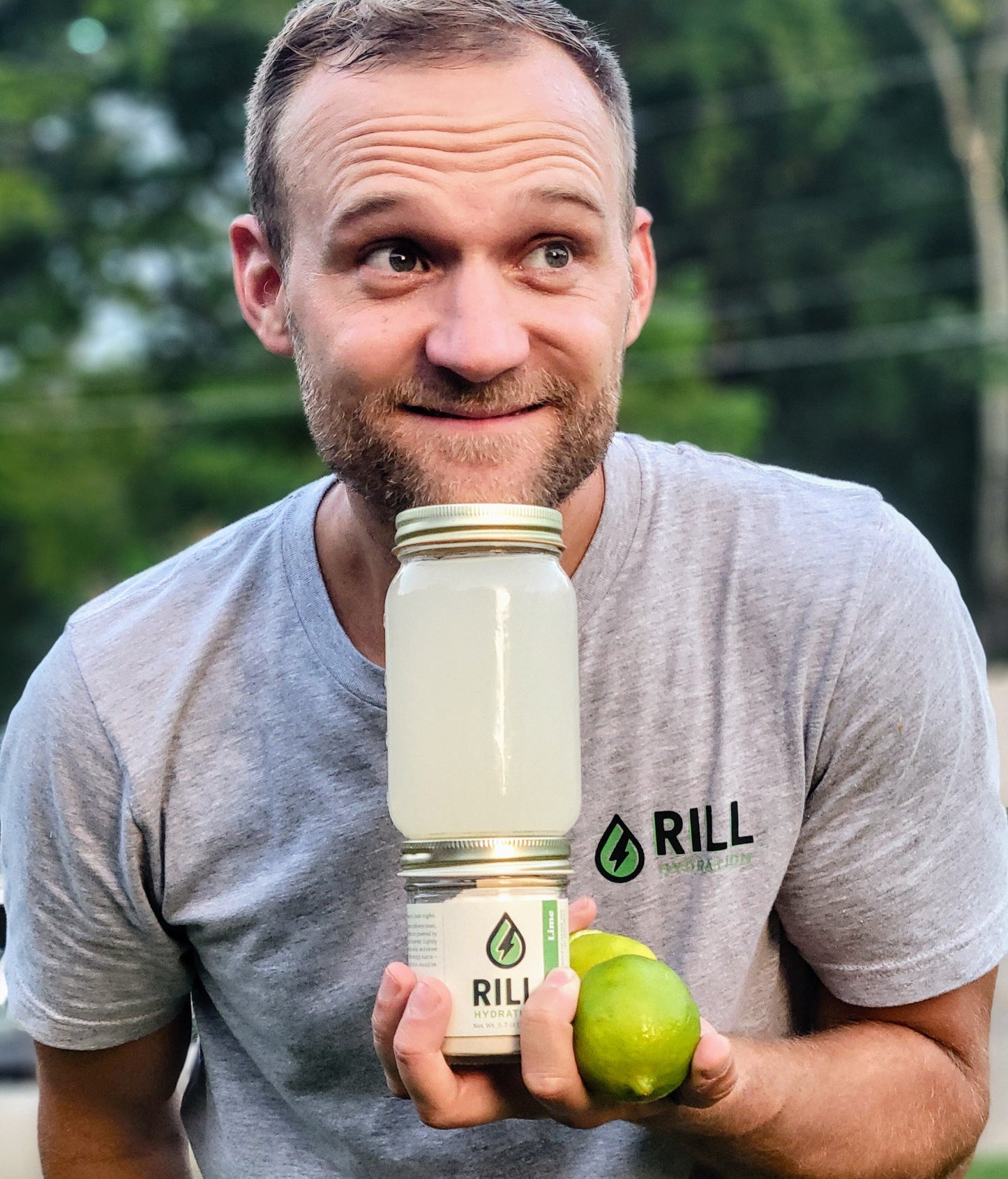 A man in a gray Rill Hydration shirt holds a jar of cloudy Lime Rill - Clean Hydration Drink Mix (30 Serving Bulk Jar) with a lime outdoors, greenery blurred in the background.