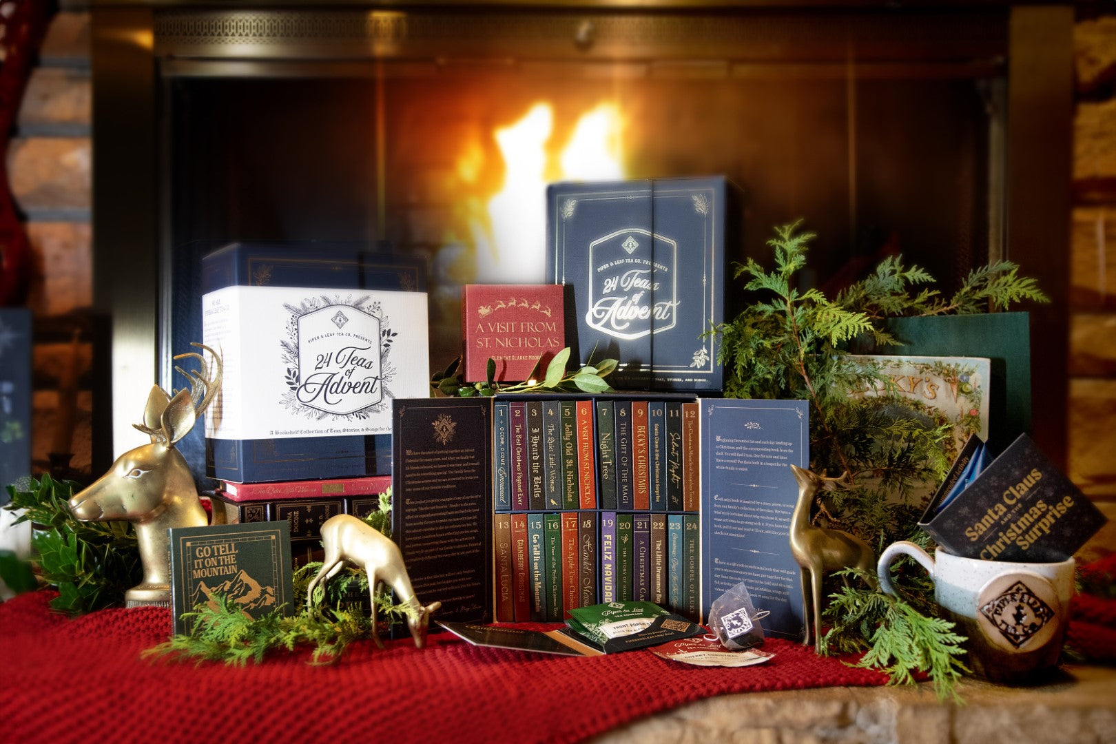 A display featuring the 24 Teas of Advent—Piper and Leaf Advent Calendar by Piper & Leaf Tea Co., along with books, greenery, holiday gift sets, and gold reindeer figurines, is arranged on a red cloth before a lit fireplace.