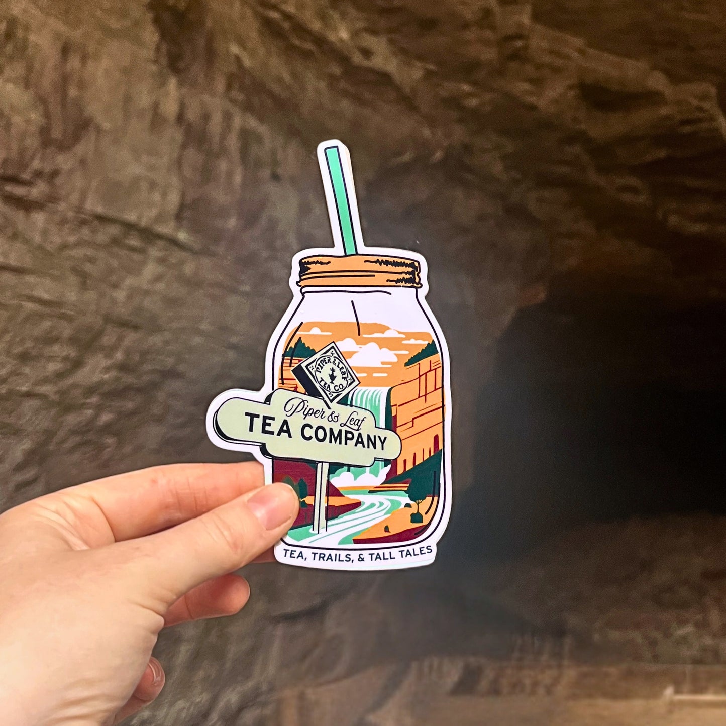 A hand holds the 'Tea, Trails & Tall Tales Adventurer Sticker' from Piper & Leaf Tea Co., showcasing a jar with a straw and landscape art. In the background, a rocky cave entrance beckons wanderlust adventurers.
