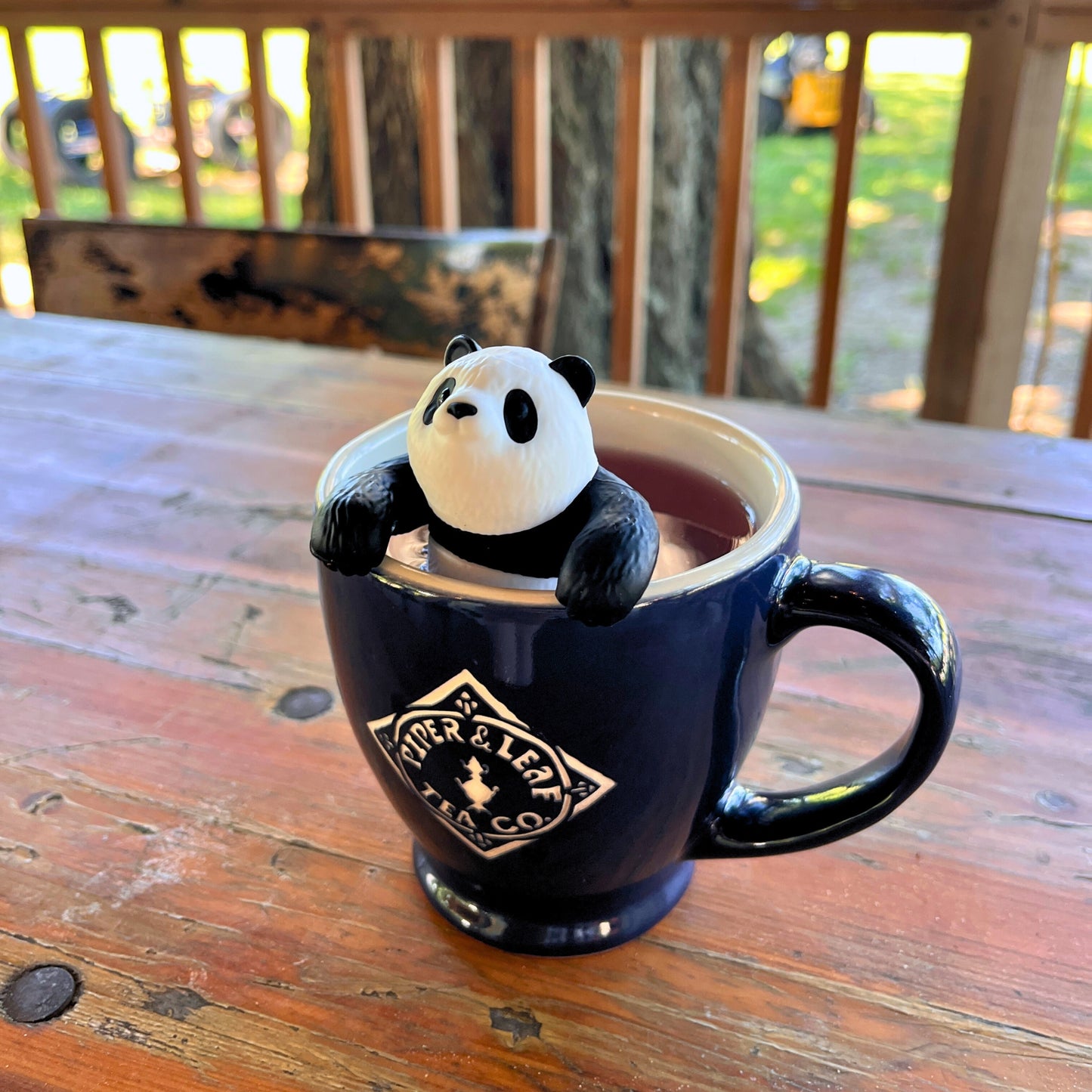 A black mug of tea with a Piper & Leaf Tea Co. BamBrew Infuser, shaped like a panda and BPA-free, designed for loose-leaf tea, sits on the rim atop a wooden outdoor table.