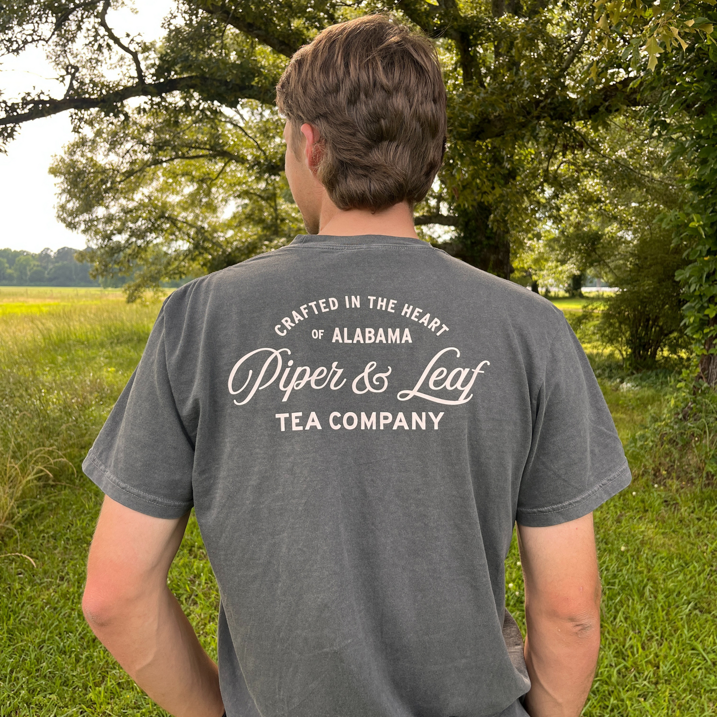 A person stands outdoors wearing the Piper & Leaf Tea Co. "Crafted in Alabama Tee," a relaxed fit, 100% cotton gray shirt featuring "Crafted in the heart of Alabama. Piper & Leaf Tea Company" on the back. Trees and grass are behind them.