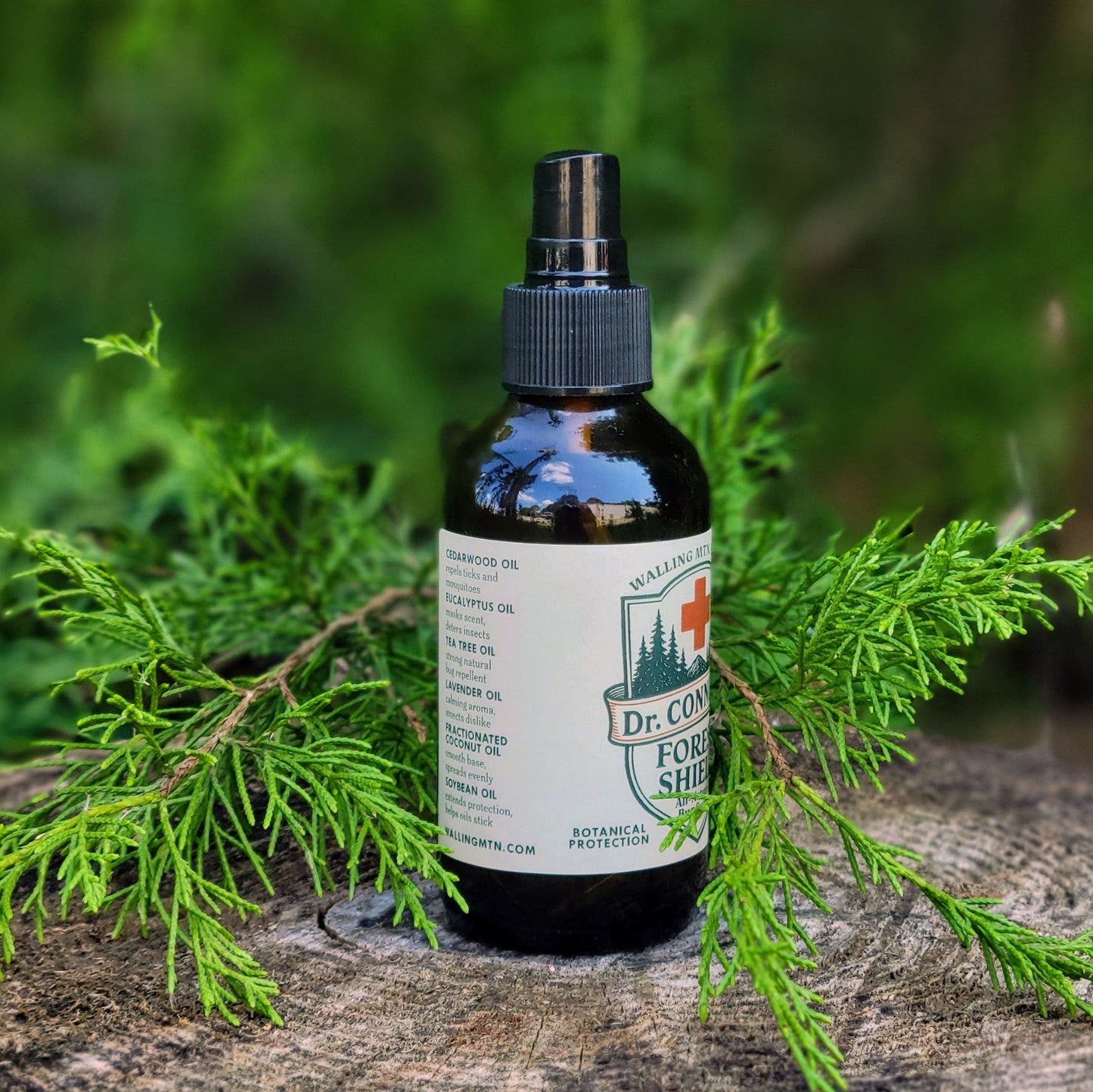 A brown spray bottle labeled "Dr. Connor's Forest Shield All-Natural Bug Spray" by Walling Mountain Farms sits on a tree stump amid cedar branches, promoting its DEET-free, natural formula for insect and mosquito protection.