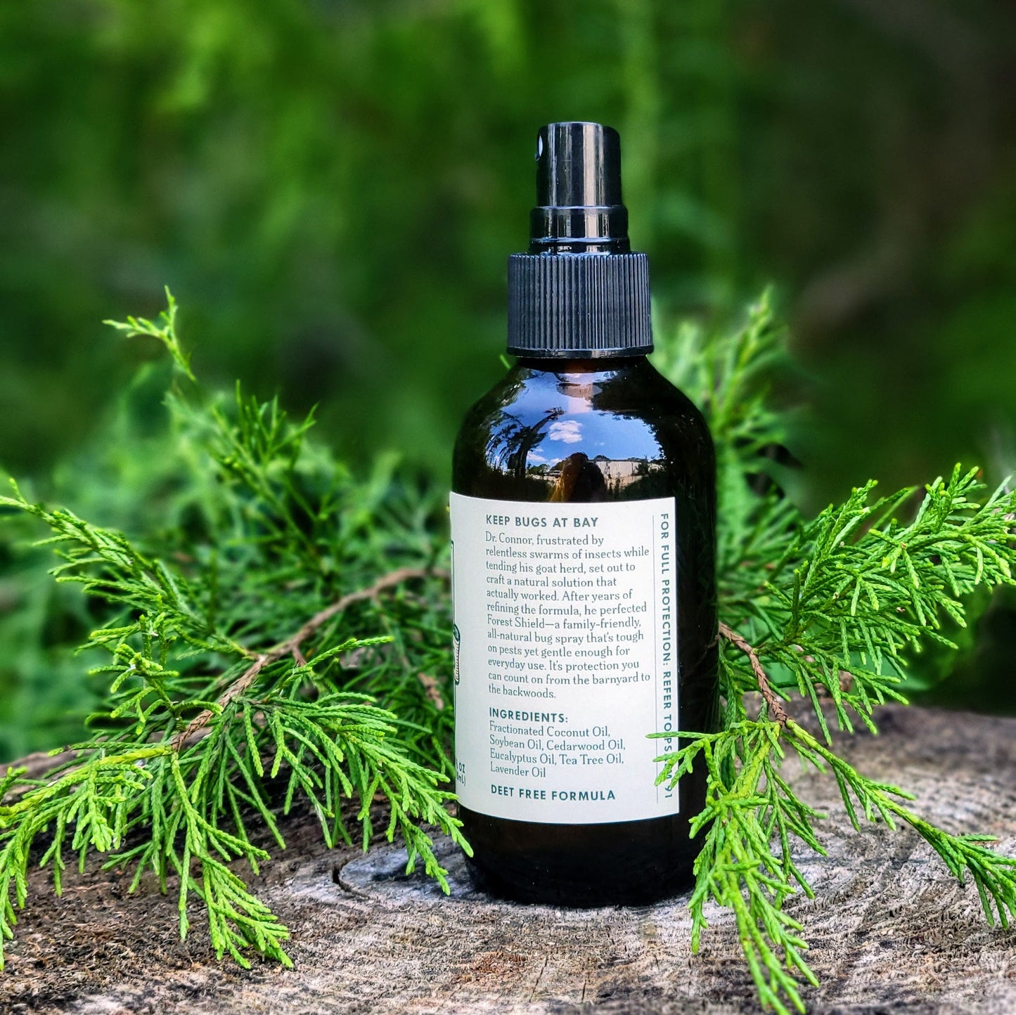 A black spray bottle of Dr. Connor's Forest Shield All-Natural Bug Spray by Walling Mountain Farms sits on a tree stump amid cedar branches, emphasizing its natural mosquito-repellent formula in a blurred outdoor setting.