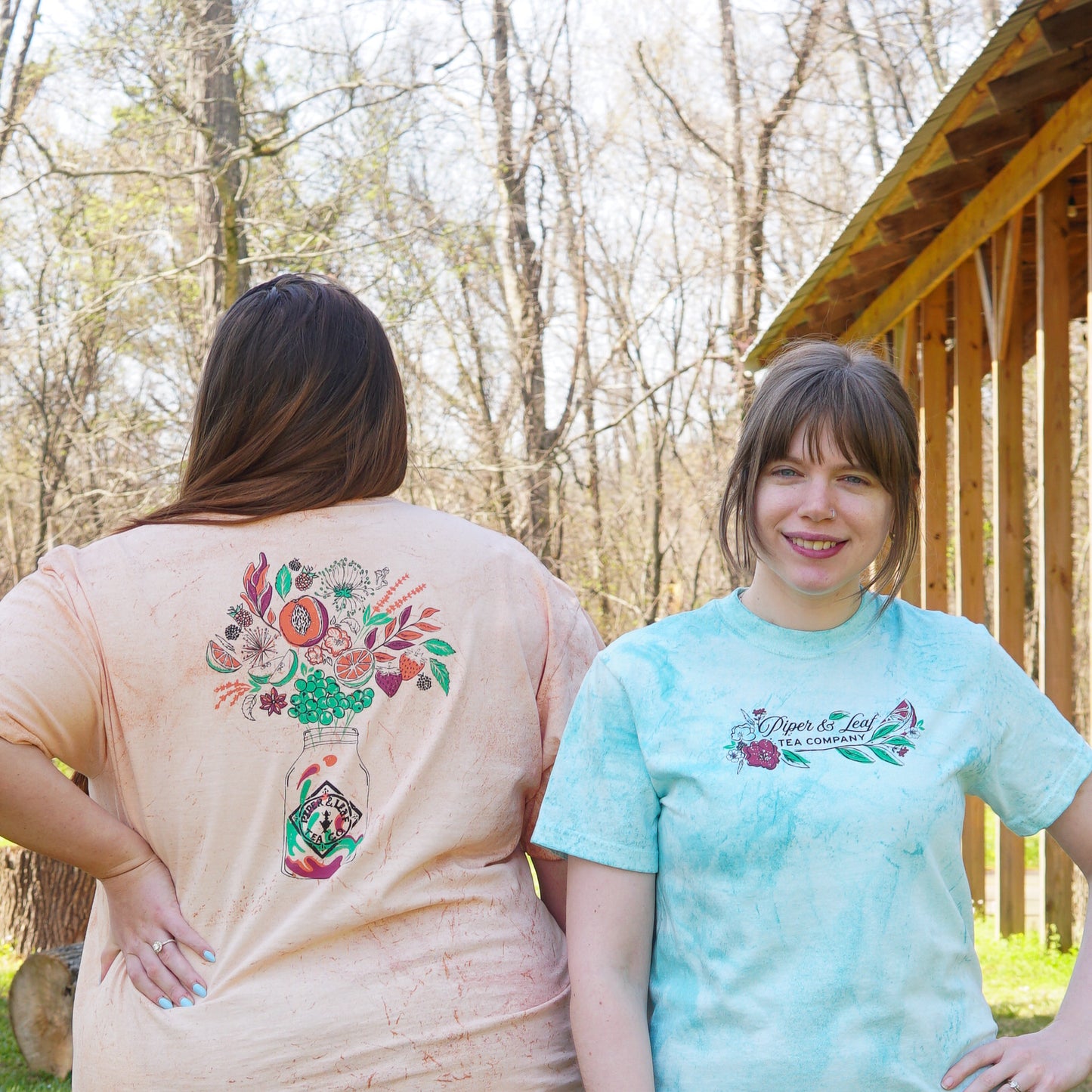 Two women stand outdoors; one faces away, displaying the floral back of a "Berries in Bloom" Short Sleeve T-shirt by Piper and Leaf Tea Co., while the other faces forward in the same shirt with its front logo visible.