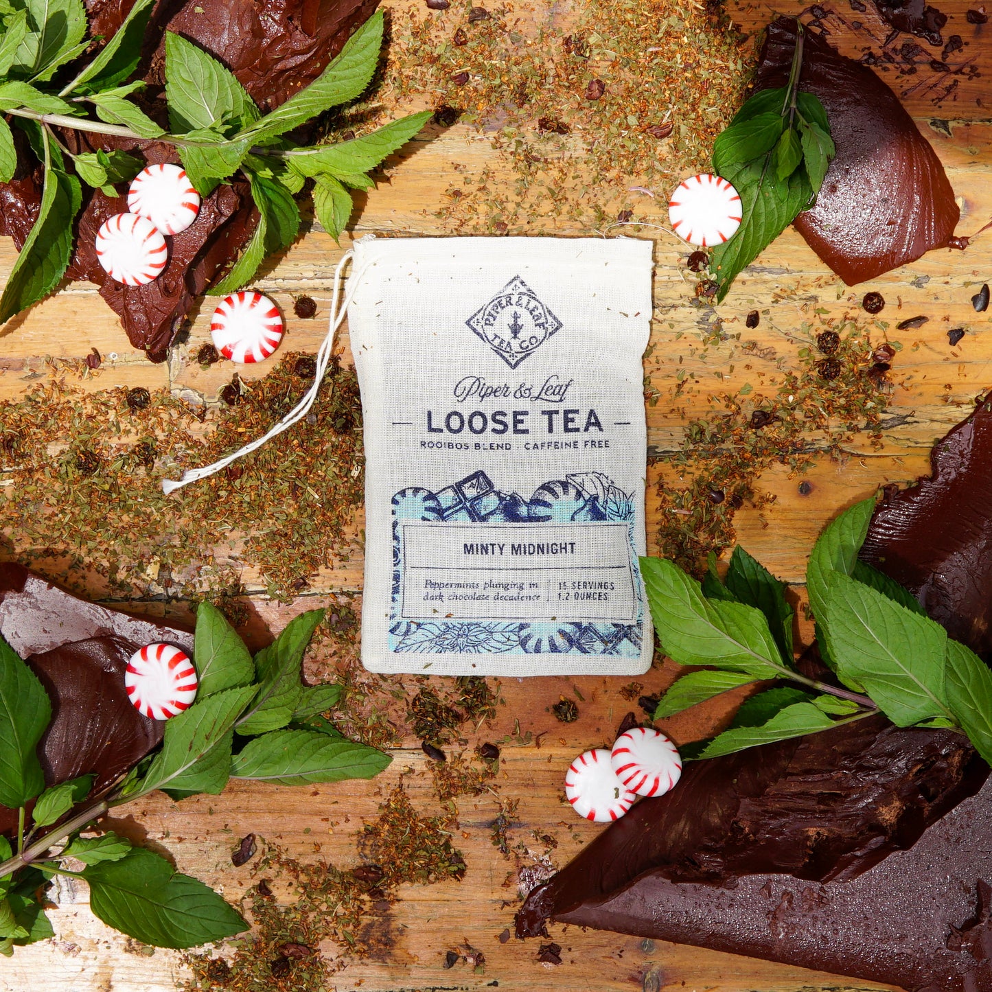 A Minty Midnight Muslin Bag of Loose Leaf Tea (15 servings) from Piper & Leaf Tea Co. is displayed with chocolate pieces, fresh mint, loose tea leaves, and peppermint candies on a wooden surface.