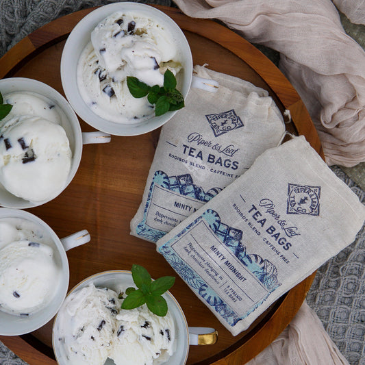 Four cups of mint chocolate ice cream with fresh mint are on a wooden tray next to two Piper & Leaf Tea Co. Minty Midnight 9ct Tea Bags in Muslin; gold spoons and a plate are placed nearby.