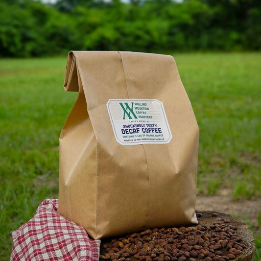A 3lb Bag of Shockingly Tasty Decaf Coffee from Walling Mtn. Coffee sits on coffee beans and a red checkered cloth outdoors, surrounded by greenery, showcasing its medium-dark roast appeal.
