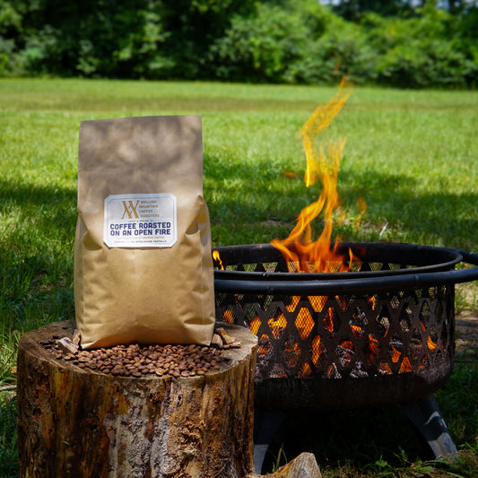 A 5lb Bag of Coffee Roasted on an Open Fire by Walling Mtn. Coffee sits on a tree stump with beans near a fire pit in a grassy outdoor setting—the perfect choice for small batch coffee fans.