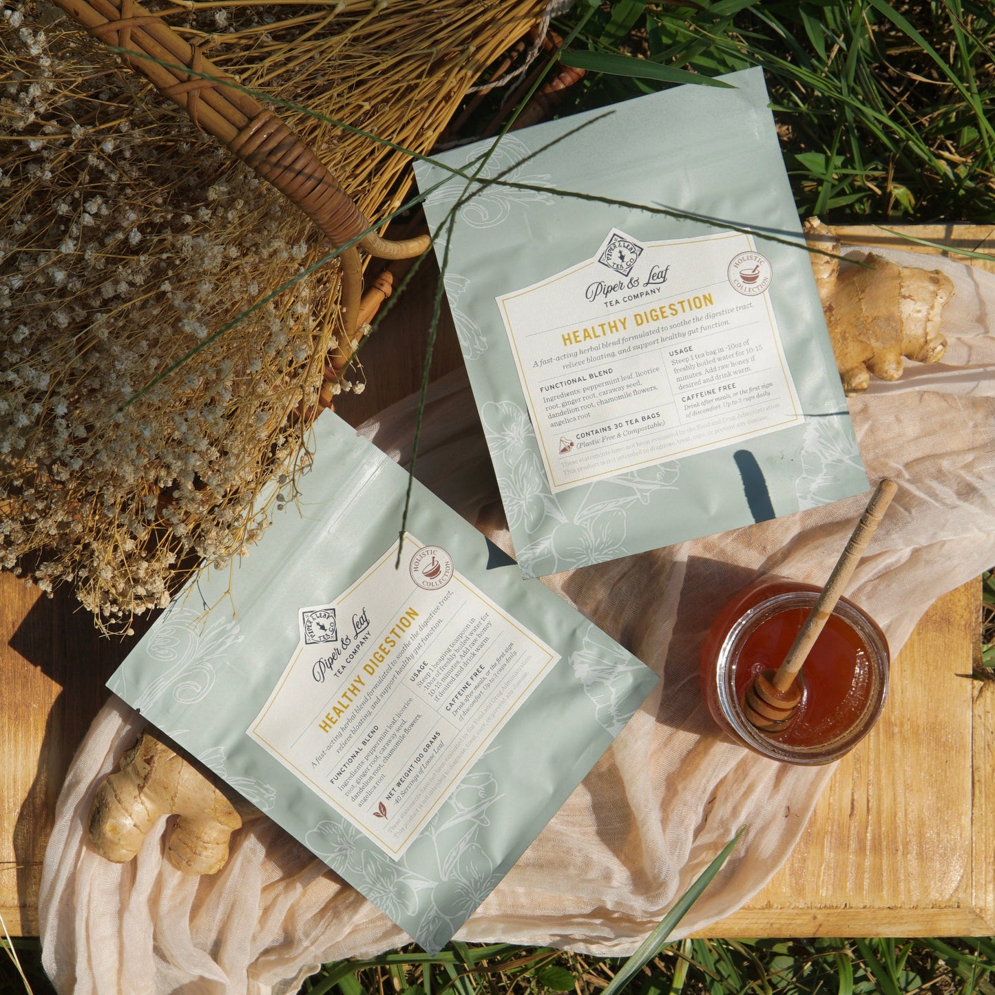 Two packets of Piper & Leaf Tea Co.’s "Healthy Digestion" tea, a soothing herbal blend for digestive support, are displayed on a wooden surface alongside ginger root, honey, dried flowers, and greenery.