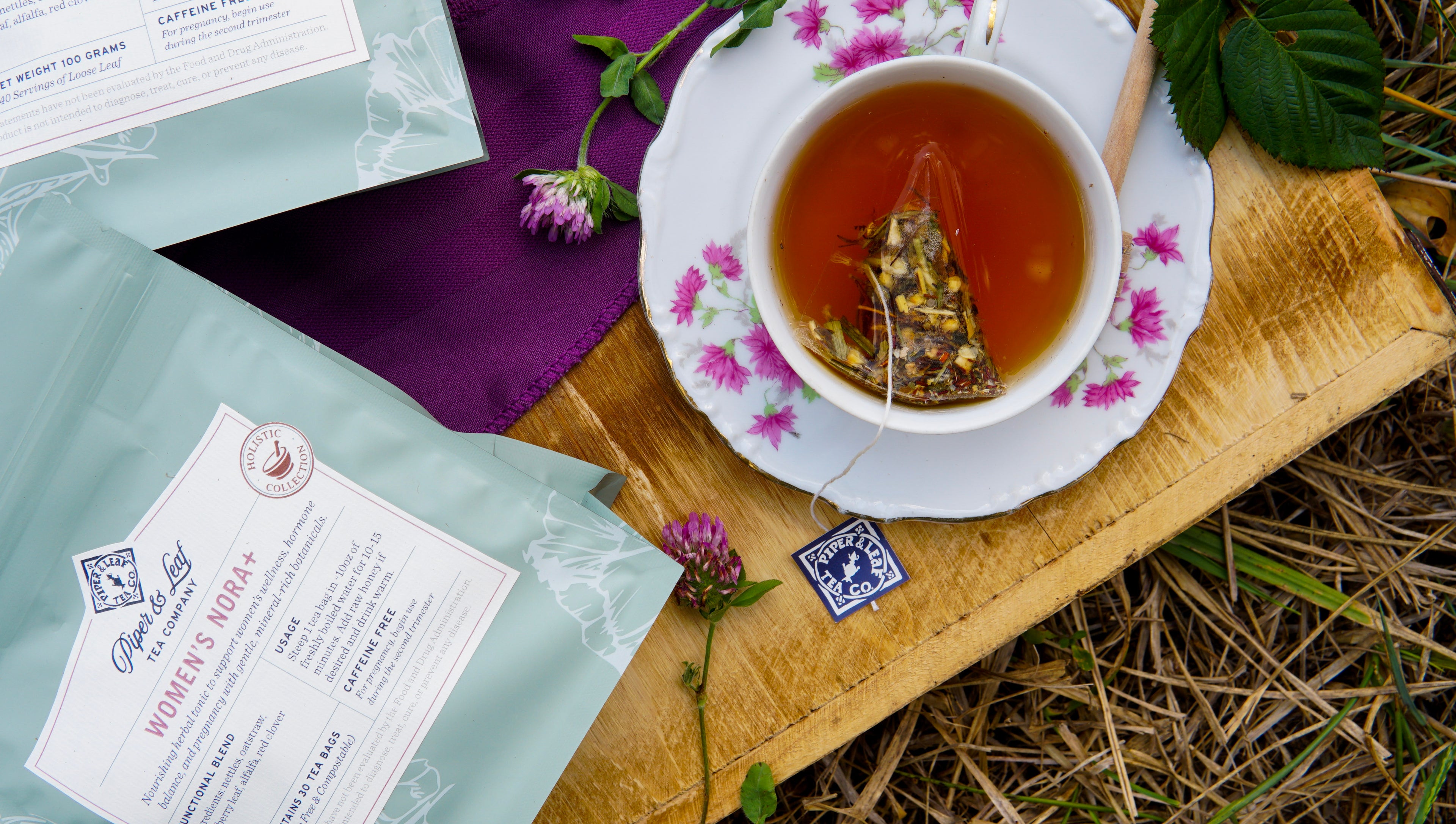 A floral teacup with herbal tea sits on a wooden board beside Piper & Leaf Tea Co.’s Women’s NORA+ packaging, clover flowers, and a purple cloth—an inviting outdoor scene inspired by women’s wellness.