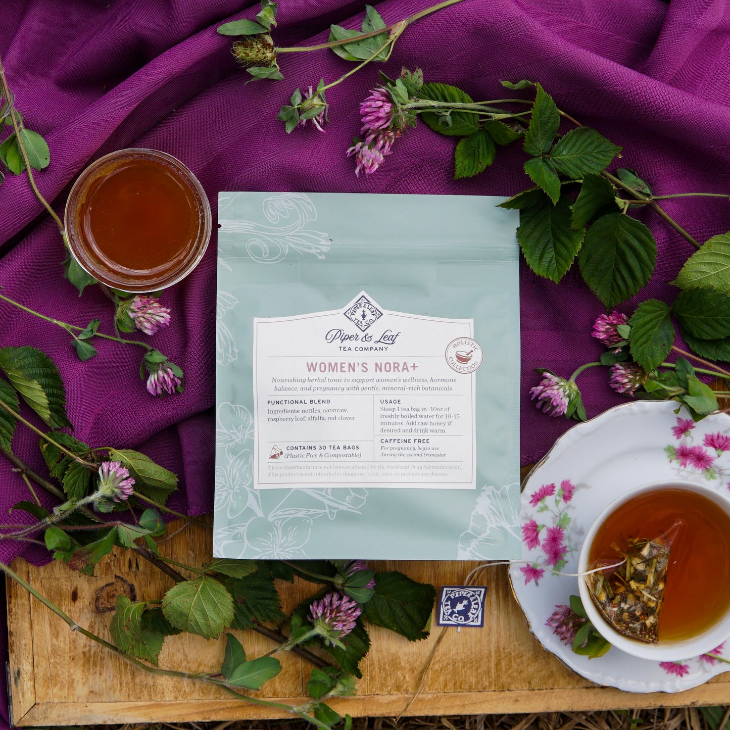 A package of Piper & Leaf Tea Co.’s Women’s NORA+ tea is displayed with a tea cup, a glass cup, and green leaves on a purple cloth atop a wooden surface.