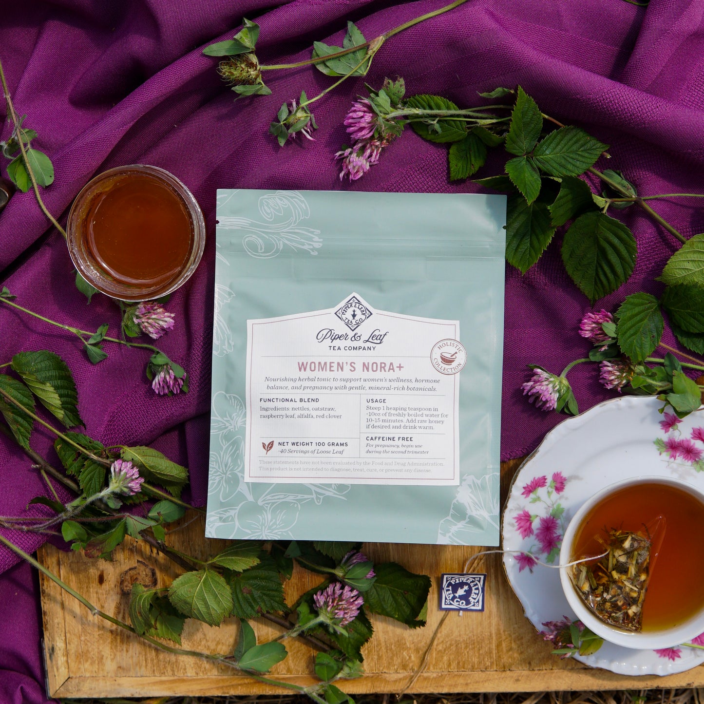 A packet of Piper & Leaf Tea Co.'s Women’s NORA+ sits on a wooden surface, surrounded by tea cups, green leaves, clover flowers, and a purple cloth—creating an inviting mood for women’s wellness and hormone balance.