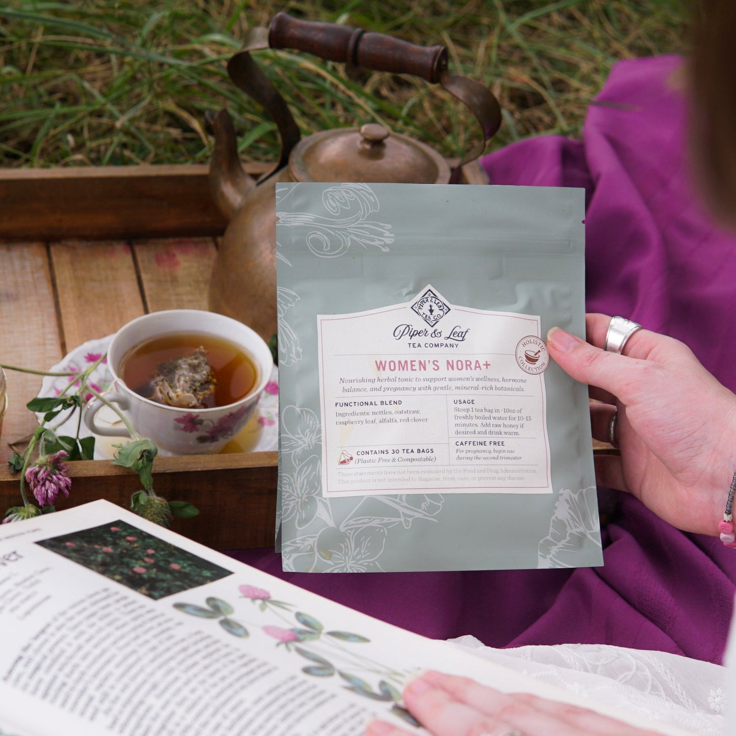 Someone holds a packet of Piper & Leaf Tea Co.’s Women’s NORA+—a tea blend for women’s wellness and hormone balance—beside a teacup, teapot, and open book on a wooden tray outdoors.
