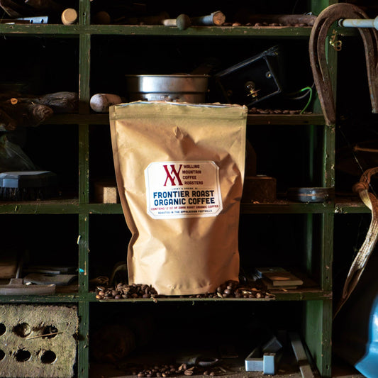 A brown 12oz bag labeled "Frontier Roast Coffee — Walling Mtn." by Walling Mtn. Coffee rests on a green shelf with tools and coffee beans, highlighting its rich blend crafted using traditional roasting methods.