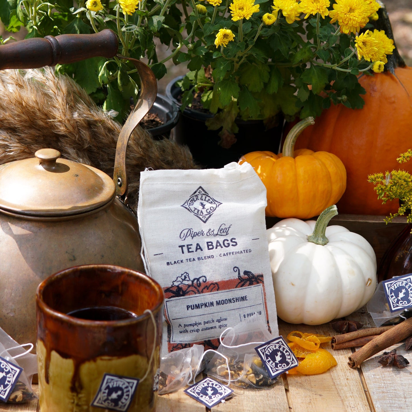 A rustic autumn scene features Piper & Leaf Tea Co.'s Pumpkin Moonshine 9ct Tea Bags in Muslin, a brass kettle, mug, yellow chrysanthemums, small pumpkins, and loose tea bags on a wooden table outdoors.