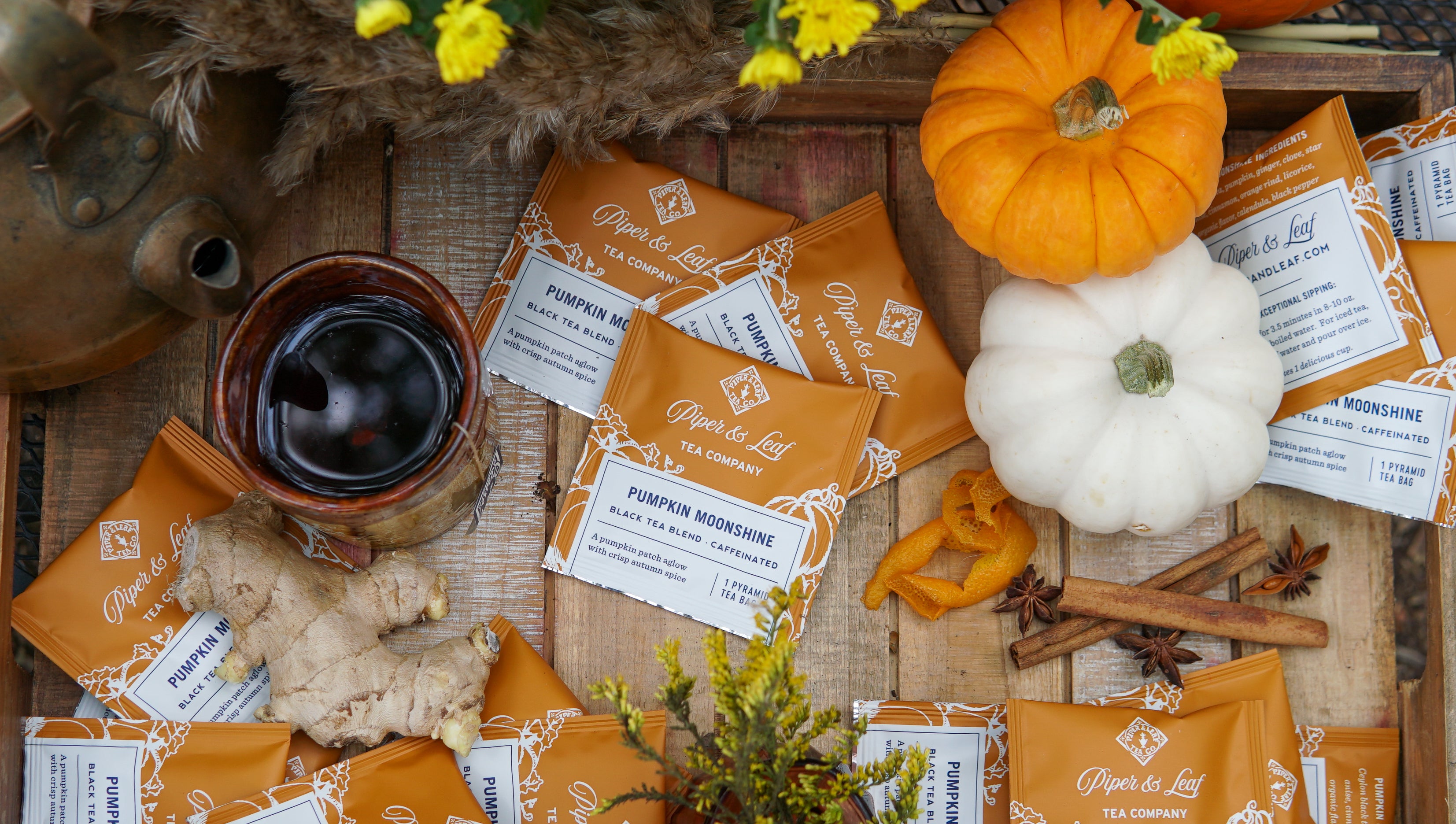 Piper & Leaf Tea Co.’s Pumpkin Moonshine Case of 50 individually wrapped tea bags is displayed with pumpkins, ginger, dried orange peel, cinnamon sticks, a teapot, and yellow flowers on a wooden surface.