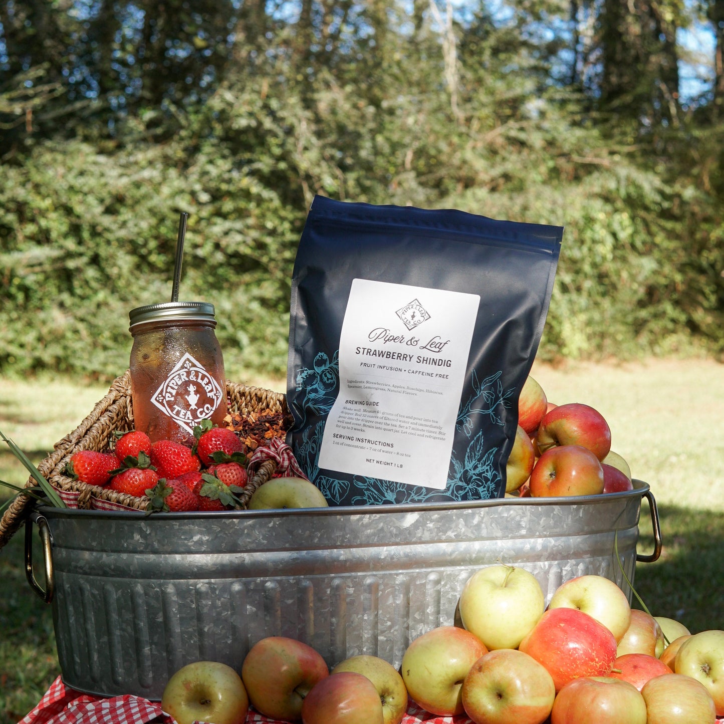 A metal tub with apples, strawberries, a mason jar of iced tea, and a Piper & Leaf Tea Co. Strawberry Shindig Pound Bag (190 servings) rests outside on a red checkered cloth.