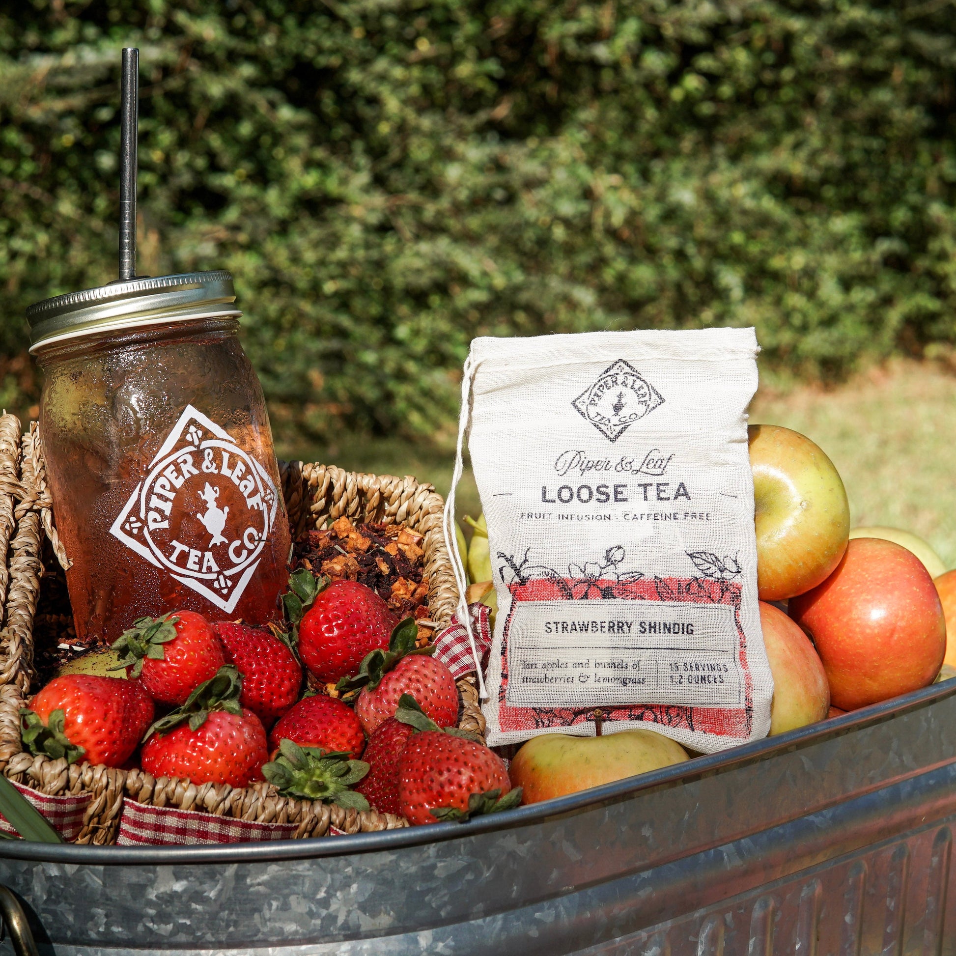 A metal tub holds baskets of strawberries, apples, iced tea with a straw, and a muslin bag labeled “Piper & Leaf Tea Co. Strawberry Shindig Loose Leaf Tea - 15 Servings.” Enjoy this caffeine-free, strawberry lemongrass blend. Bushes are in the background.