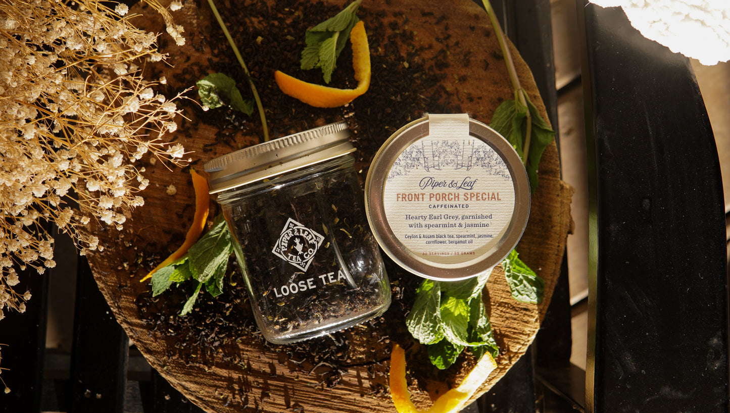 A Piper & Leaf Tea Co. Front Porch Special Glass Jar of Loose leaf Tea (30 servings), on a wooden board with orange peels, mint leaves, and dried flowers—55g per jar for the perfect flavorful brew.