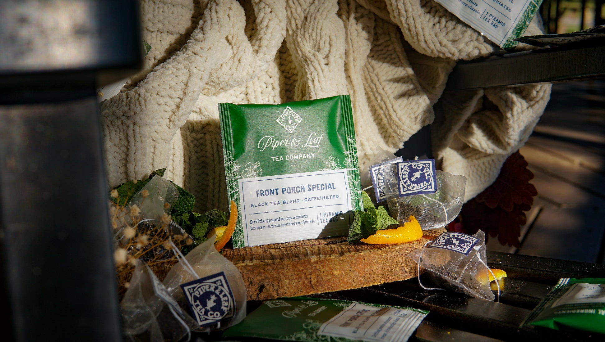 A green packet of Piper & Leaf Tea Co. "Front Porch Special" Case of Tea Bag Envelopes (50ct) sits on a bench, surrounded by tea sachets, fallen leaves, and a cozy knit blanket.
