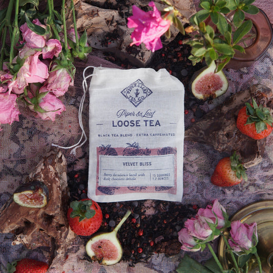 A Velvet Bliss Muslin Bag of Loose Leaf Tea (15 servings) from Piper & Leaf Tea Co. sits on a lace cloth, surrounded by strawberries, pink flowers, tea leaves, and chocolate pieces, inspired by chocolate-covered strawberry tea.