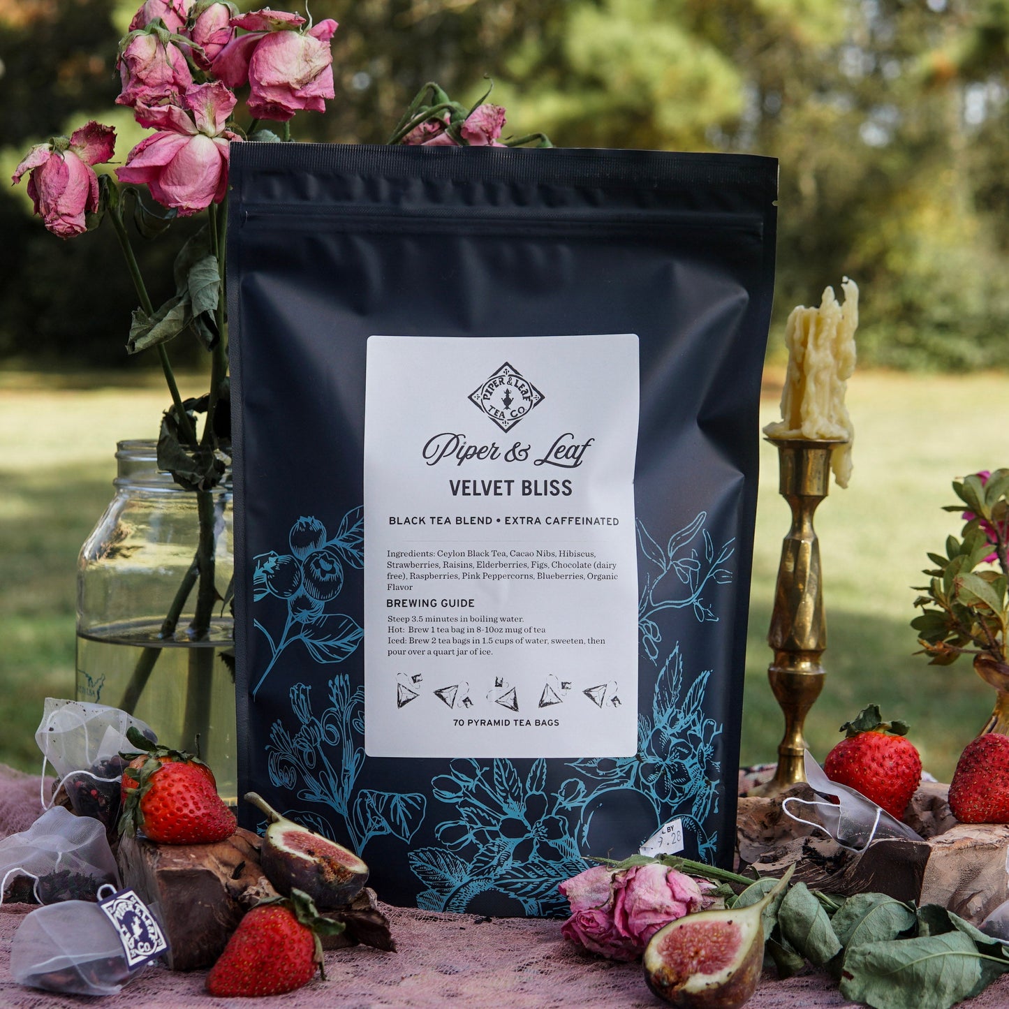 A bag of Piper & Leaf Tea Co. Velvet Bliss Bulk Sachets – 70ct Tea Bags rests outdoors beside strawberries, dried roses, tea sachets, and a melted candle, evoking the aroma of creamy rich chocolate tea.