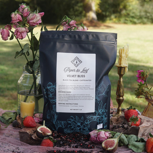 A Piper & Leaf Tea Co. Velvet Bliss Pound Bag (190 servings) is displayed outdoors on a cloth with dried roses, strawberries, and decorative candles.