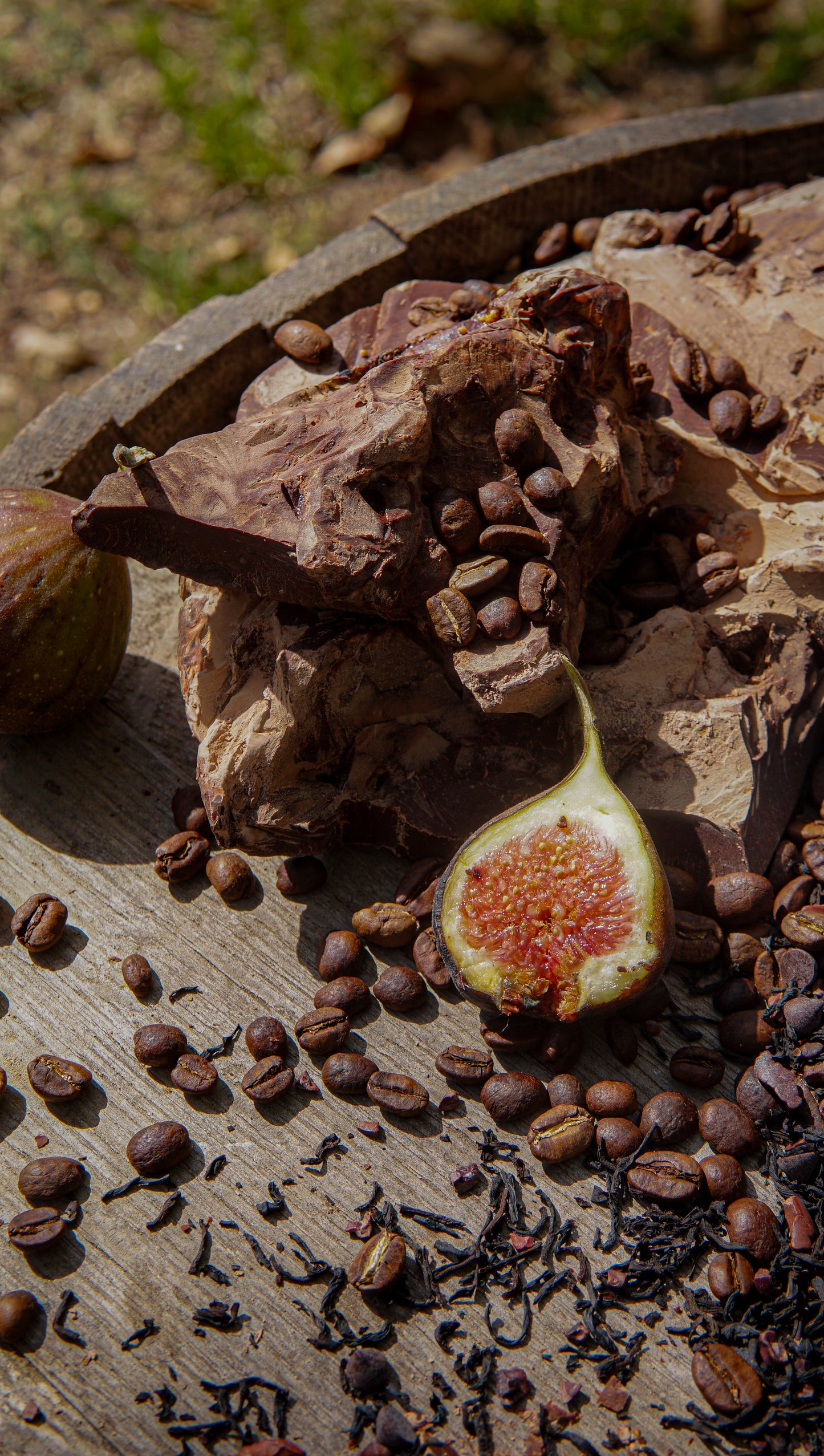 A fig half, coffee beans, chocolate chunks, and Piper & Leaf Tea Co.'s Chocola-Tea Muslin Bag of Loose Leaf Tea (15 servings) are artfully arranged on a wooden surface outdoors.
