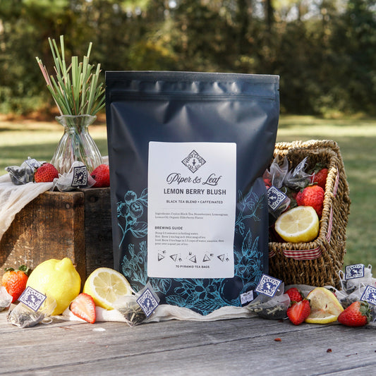 A box of Piper & Leaf Tea Co.'s Lemon Berry Blush Bulk Sachets (70ct tea bags) sits outdoors on a wooden surface, surrounded by lemons, strawberries, a wicker basket, and several tea bags.