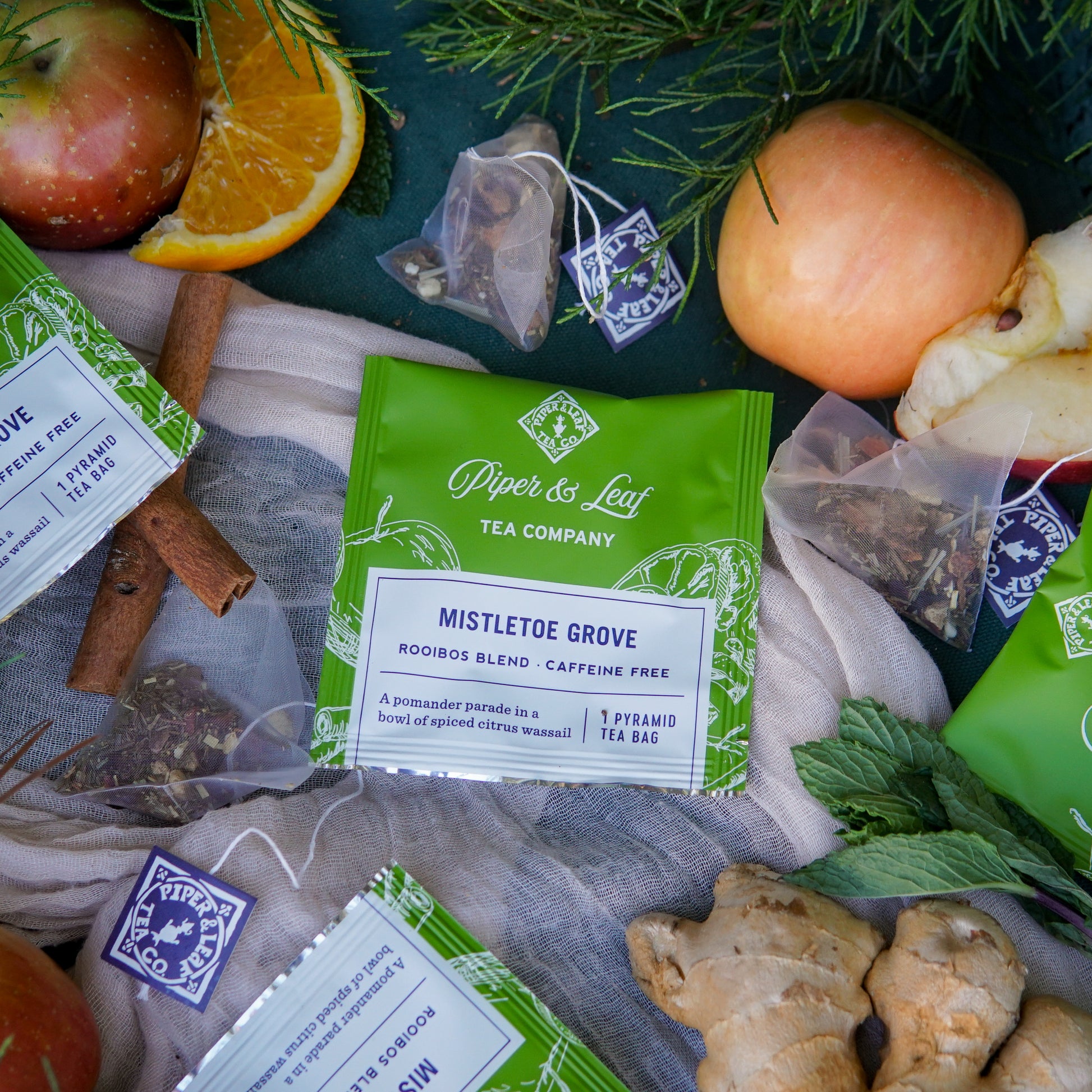 A case of 50 individually wrapped Mistletoe Grove tea bags by Piper & Leaf Tea Co., surrounded by ginger, apples, orange slices, cinnamon sticks, mint leaves, and loose tea bags.