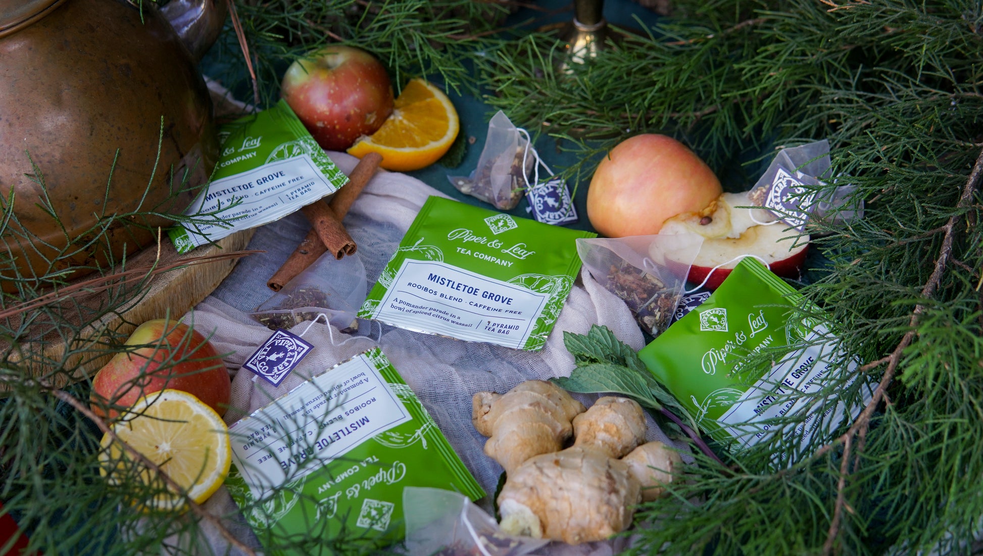 A case of Piper & Leaf Tea Co. Mistletoe Grove individually wrapped tea bags (50ct) is arranged on a cloth with fresh ginger, apples, lemon, orange slices, cinnamon sticks, and pine branches.