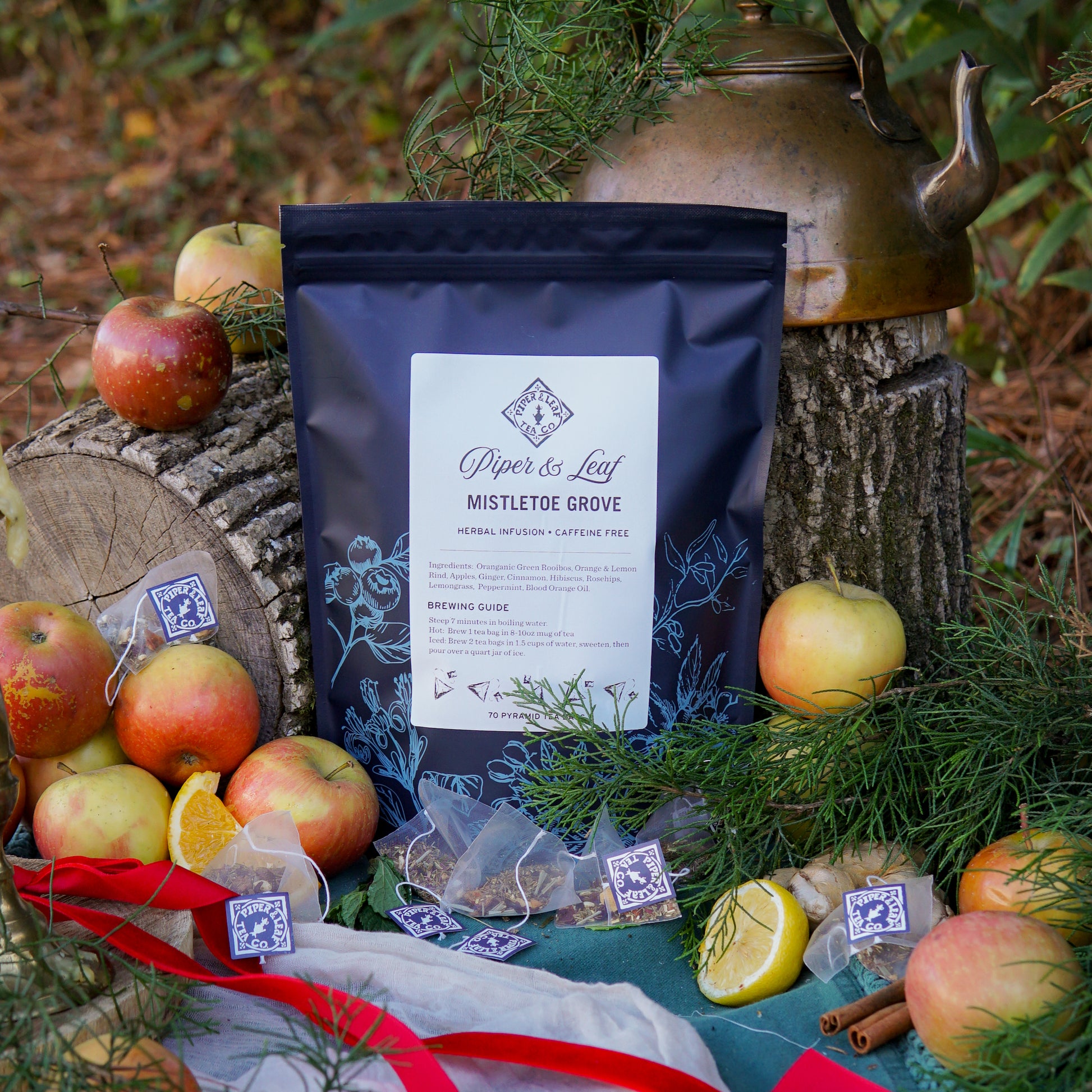 A box of Piper & Leaf Tea Co. Mistletoe Grove Bulk Sachets (70ct tea bags) is displayed outdoors with apples, orange slices, herbs, tea bags, and a brass kettle arranged on tree stumps and grass.