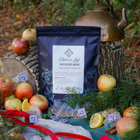 A box of Piper & Leaf Tea Co. Mistletoe Grove Bulk Sachets (70ct tea bags) is displayed outdoors with apples, orange slices, herbs, tea bags, and a brass kettle arranged on tree stumps and grass.