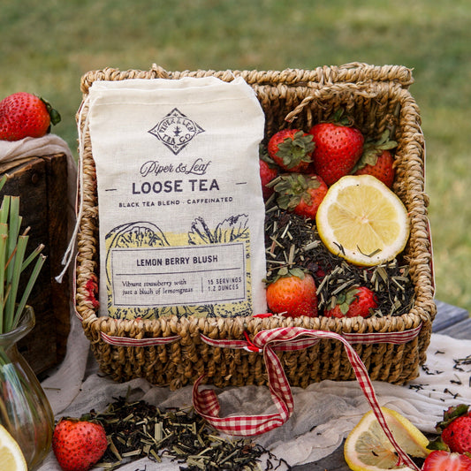 A basket featuring a Piper & Leaf Tea Co. Lemon Berry Blush Muslin Bag of Loose Leaf Tea (15 servings), surrounded by loose tea leaves, strawberries, lemon slices, and a red-checked ribbon.