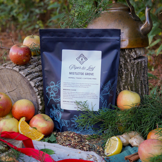 A Piper & Leaf Tea Co. Mistletoe Grove Pound Bag (190 servings) is shown outdoors with apples, oranges, fir branches, a brass kettle, and a log—ideal for cozy holiday tea by the fire.