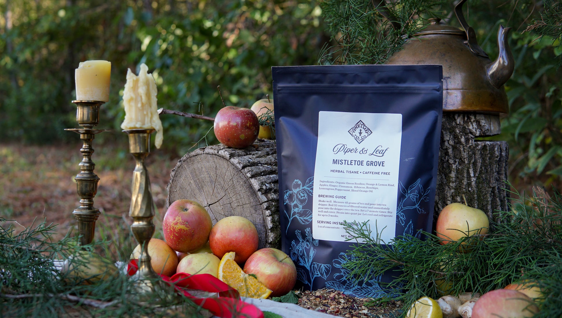 The Piper & Leaf Tea Co. Mistletoe Grove Pound Bag (190 servings) is featured outdoors with apples, oranges, a brass teapot, candlesticks, and greenery arranged on a log and the forest floor.